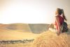 Woman with a backpack sitting on a hay bale, enjoying a serene sunset over a vast countryside field.