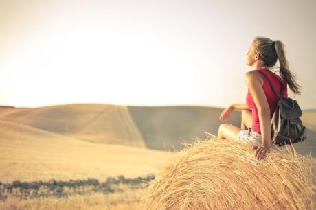 Woman with a backpack sitting on a hay bale, enjoying a serene sunset over a vast countryside field.